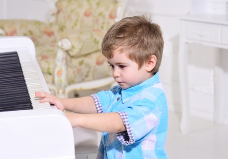 Child sit near piano keyboard, white background. Kid spend leisure near musical instrument. Boy cute and adorable puts finger on keyboard of piano. Elite childhood concept.の写真素材