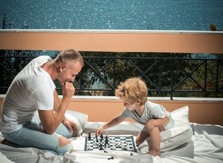 Father and little son play chess on balcony, nature and sea on background. Dad with child play intellectual game. Smart toddler concept. Parent play chess with kid on terrace on sunny day.の写真素材