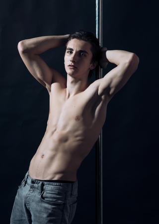 Sexy, attractive guy lean on metallic pole. Macho with naked chest, athlete, sportsman, dancer performing pole dancing moves. Sexy dancer concept. Man on pensive face with nude torso, black backgroundの写真素材