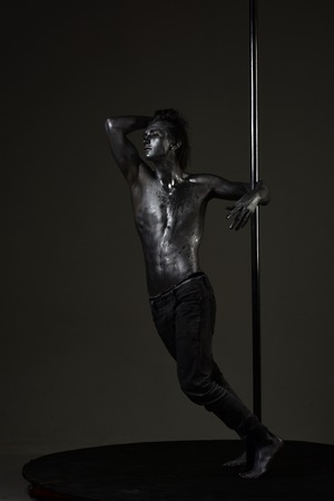 Pole dance man on pensive face stand near metallic pole, dark background. Guy with nude torso performing pole dancing moves on pole.の写真素材