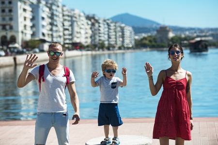 Mother and father and their child waving hands. Happy family spend time together, waving hands, sea and urban background. Parents with son stand on seafront. Family vacation concept.の写真素材