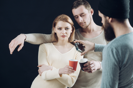 Men, woman on pensive faces, black background. Men pouring womans cup with alcohol out of flask. Get drunk concept. Company of cheerful friends speaks and drinks. Communication, friendship, fun.の写真素材