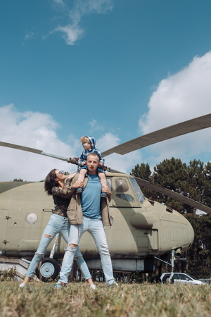 Happy family spend time together, on excursion, helicopter on background, sunny day. Family leisure concept. Mother and father and child walking in aviation museum outdoors. Dad carries son on neck.の写真素材