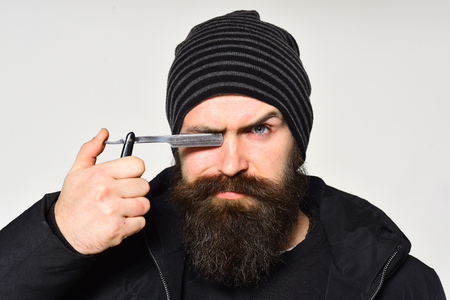 Barber, hipster barbershop assistant with great beard holds razor near eye