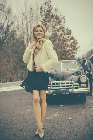 Woman walks on road. Couple in love on romantic date. Retro collection car and auto repair by mechanic driver. Bearded man and sexy woman in fur coat. Travel and business trip or hitch hikingの写真素材
