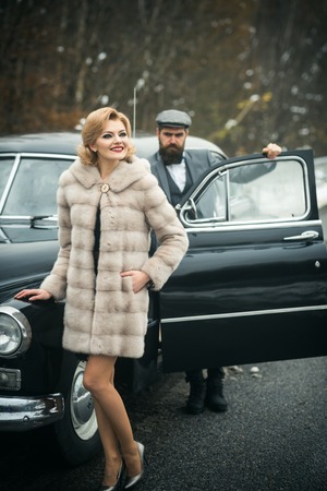 Bearded man and sexy woman in fur coat. Escort of girl by security. Retro collection car and auto...