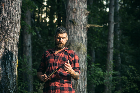 Brutal lumberjack looking at small sharp spade in his hands. Bearded man wandering in forest in the evening. Lone hiker in woods at sunset, summer night camping.の写真素材