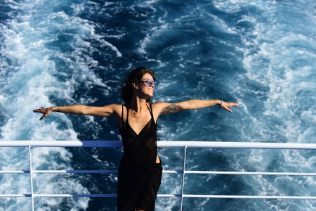 Attractive girl feels free and enjoy summer vacation. Freedom concept. Sexy lady stands on deck of cruise liner with sea waves on background. Woman in dress travelling on cruise ship on sunny day.の写真素材