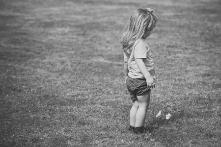 Boy small cute kid with blond long hair in green shirt and blue shorts standing and looking at little white flower on background of grass in sunny day.の写真素材