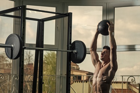 Sport and gym concept. Sportsman, athlete , muscular macho does exercise with medicine ball, window and barbell on background. Man with nude torso in gym enjoy training, trx.の写真素材
