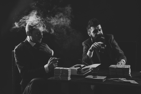 Illegal business concept. Businessmen discussing illegal deal while drinking and smoking, dark background. Company engaged in illegal business. Men sitting at table with piles of money and typewriter.の写真素材