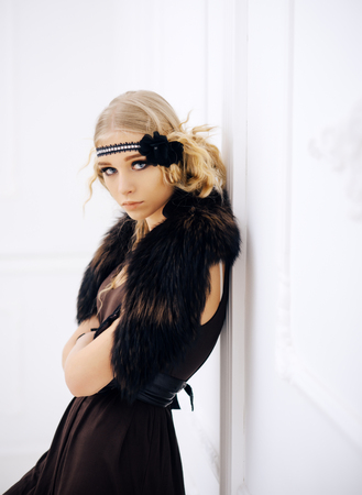 Young movie star of roaring twenties posing isolated on white background. Blond girl in headband with flower and fur collar leaning on wall.の写真素材