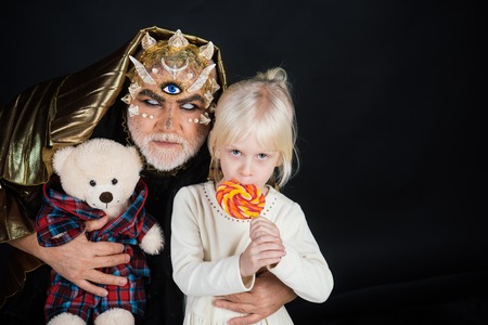 Senior man with white beard dressed like monster telling story to little girl. Fairytale concept. Man with thorns or wart with child sitting on his knees. Demon with golden hood on black background.の写真素材