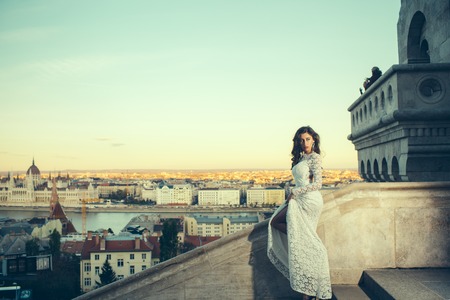 Girl with glamour look. Woman in white wedding dress on city view, fashion. Sensual woman with long hair on balcony, beauty. Bride with makeup. Fashion model style and hairstyleの写真素材