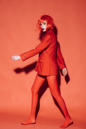 Fashion concept. Lady with dark lips looking at camera and walks. Woman with makeup and red wig posing in total red outfit. Girl on mysterious face in red formal jacket, red background.の写真素材