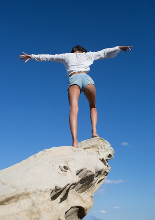 Rear view of a beautiful, brunette young girl with raised hands, looking at ocean. Freedom concept, holiday on beach with clear sky background.の写真素材