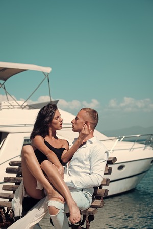 Young passionate couple sit on bench at dock near boat and hugs, sunny summer day. Woman and man in fashionable clothes full of desire near luxury yacht. Honeymoon concept. Couple in love on honeymoonの写真素材