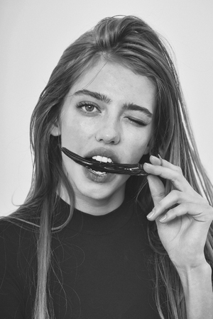 sexy pretty girl or beautiful woman with long, blond hair, hairstyle and adorable winking face in black sweater holds red chilly pepper in lips, mouth isolated on white backgroundの写真素材