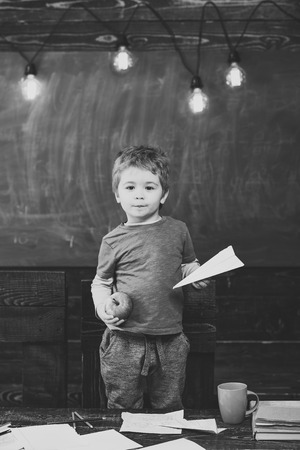 Little boy holding paper plane and apple in his hands. Schoolboy in blue T-shirt standing behind the table. Educational game concept.の写真素材