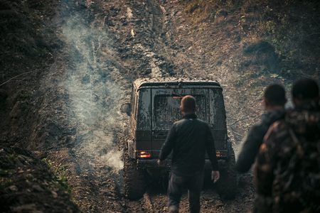 Company of three men looks at suv drives with clouds of smoke. Car covered with mud on path with deep rut. Offroad car with dirt on background. Extreme entertainment concept.の写真素材