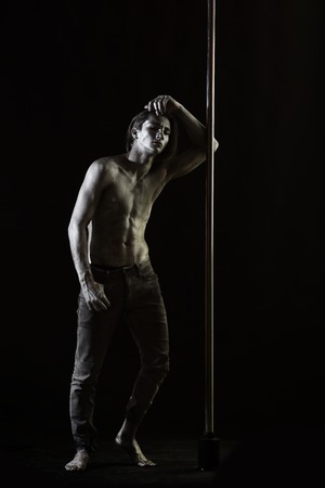 Tired dancer concept. Guy lean on metallic pole. Man with naked chest, athlete, sportsman performing pole dancing moves. Man with nude torso covered with shimmering silver paint, dark background.の写真素材