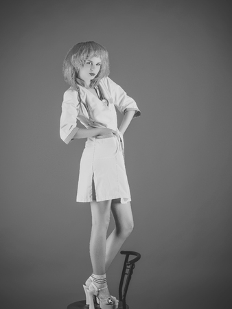 Woman in doctor uniform with stethoscope standing on chair. Nurse girl in red wig and shoes with high heels on blue background. Health and medical care concept.の写真素材