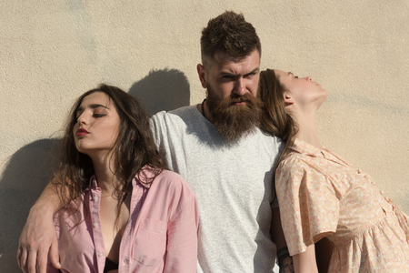 Man with beard hugs two ladies on hot sunny day. Threesome suffers of heat on summer day, wall on background. Girls turned to opposite sides while man hugs them. Love triangle concept.の写真素材