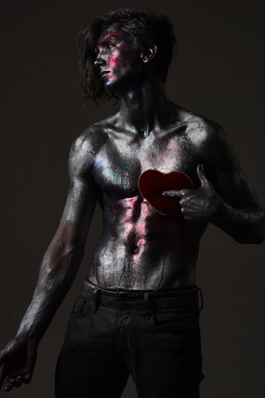 Man with nude torso holds red plush soft heart toy on chest, dark background. Loneliness and brake up concept. Guy on sad face with make up, covered with shimmering silver paint and colorful glitters.の写真素材