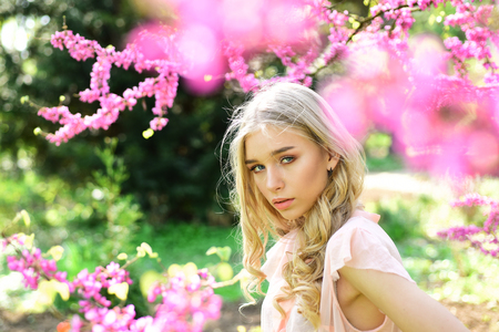 Beautiful girl standing under gentle tree with tiny hot pink blossom. Charming blond young woman breathing fresh aroma of blooming flowers outdoors, spring time concept.の写真素材