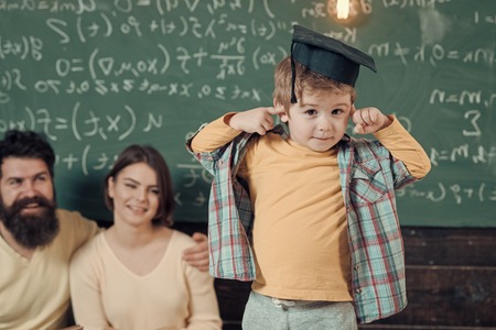 Happy kid having fun. Parents listening their son, chalkboard on background. Boy presenting his knowledge to mom and dad. Wunderkind concept. Smart child, wunderkind in graduate cap pointing at his head.の写真素材