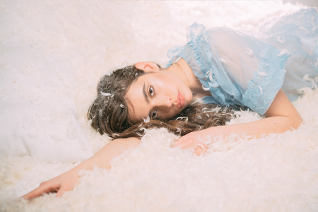 Beautiful brunette teenage girl lying on bed after pillow fight, tiny feathers flowing in air. Tired, drowsy female with wavy hair falling asleep.の写真素材