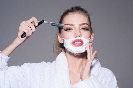 Lady play with sharp blade of straight razor. Girl on dreamy face wears bathrobe, grey background. Woman with face covered with foam holds straight razor in hand. Barber and shaving concept.の写真素材