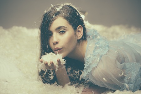 Beautiful girl in lace nightgown with fluff in hair blowing handful of feathers in air. Lovely nymph lying on downy bed. Fairy tale, fantasy and dream land conceptの写真素材