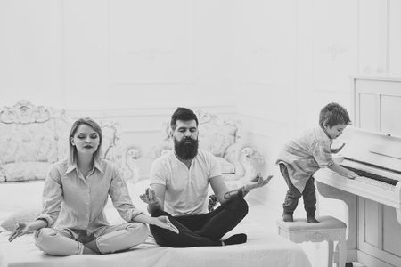 Mom and dad try to keep calm. Meditation concept. Young parents meditating, while their little son playing piano, annoying parents. Couple do yoga exercises in bed. Kid annoyed parents with noiseの写真素材
