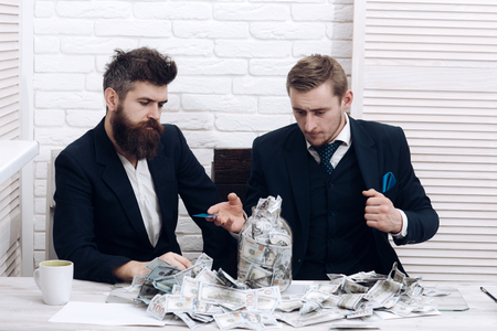 Business partners, businessmen at meeting in office. Cash issues concept. Colleagues collecting money in jar instead of bank account. Manager with beard and colleague with jar of cashの写真素材