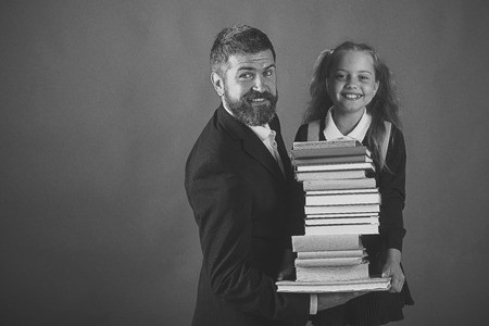 It's time to go to school. Girl in school uniform and bearded man in suit. Father and schoolgirl with happy faces on dark green background. Home schooling and back to school concept. Kid and dad hold huge pile of booksの写真素材
