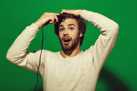 Men's haircut. surprised man shaving with electric razor or shaver, trimmer with long hair and beard on face in white underwear on green background, morning and everyday life, barbershop and fashionの写真素材