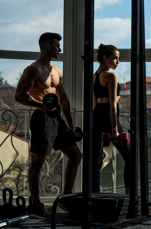 Man and woman in sportswear in gym, window on background. Couple does physical exercises with dumbbells together. Athletes, fitness trainers muscular and strong at gym. Sport and gym concept.の写真素材