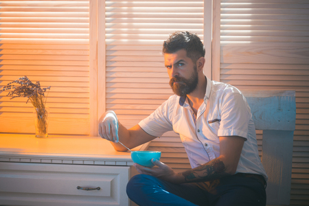 morning breakfast of serious bearded man eating from plate at window. hipster man with long beard. healthy food and snack. hungry bearded man hold breakfast in kitchen. junk food or bowl with cerealsの写真素材