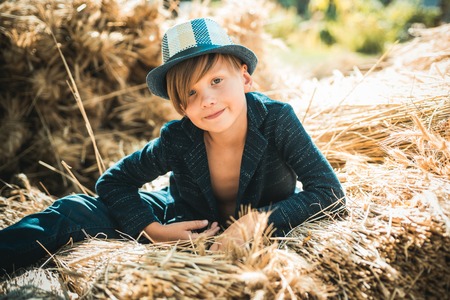 Cute little child boy holding gold leaf on farm village background. Kid boy lies on the hay. Boy in the hat are preparing for autumn sunny day.の写真素材