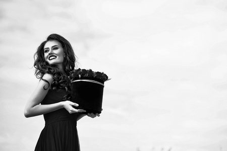 young pretty sexy woman or girl with cute smiling face and long brunette hair has fashionable makeup with red lipstick and dress holds rose flowers bouquet in box on blue sky background, copy spaceの写真素材