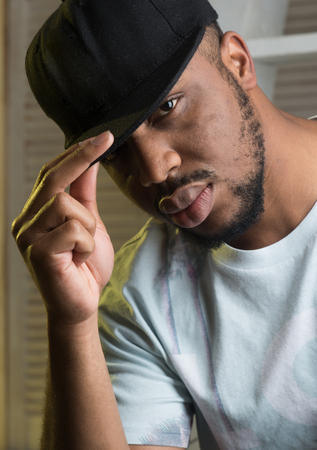 Rapper culture concept. Black macho with beard wears rapper cap and looks handsome. Man on calm face with sensual big lips, close up. Man with beard and mustache indoor, jalousie backgroundの写真素材