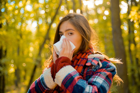 Woman with napkin sneezing in the yellow park. Sick people has runny nose. Allergy season. Woman makes a cure for the common cold. Woman makes a cure for the common cold in autumn park.の写真素材