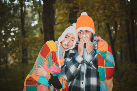 Showing sick couple sneezing at autumn park. Girl with handkerchief and sneezing boy in autumn park.の写真素材