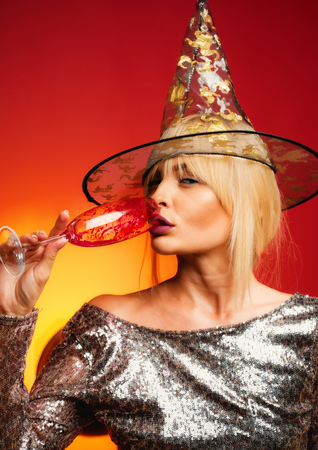 Pretty young woman in witch costume. Portrait of gorgeous blonde witch in brilliant dress and hat with glass over orange background. Beautiful young surprised woman in witches hat drinks champagne.の写真素材