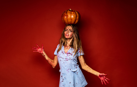 Pretty young blonde woman clothed in dress with pumpkins. Sexy zombie girl holding a pumpkin over his head. Model with bloody hands and blood on face. Halloween make-up beautiful woman.の写真素材