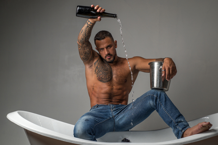 Wine festival concept. Champagne or wine celebration. Handsome brutal man pouring champagne in the bath on gray background.の写真素材