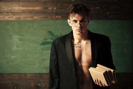 Sexy teacher concept. Teacher on pensive face, muscular figure, holds books in hand. Man with muscular torso, six packs, wears classic jacket and eyeglasses, looks attractive, chalkboard on backgroundの写真素材