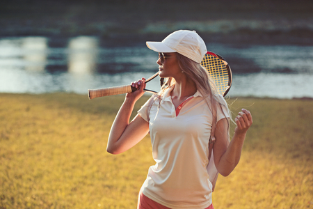 Sportswoman with tennis racket at river. Fashion woman in tennis outfit on summer landscape. Woman in cap on sunny outdoor. Sport activity and energy concept.の写真素材