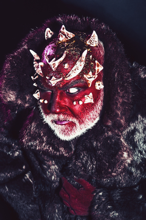 Senior man with white beard dressed like monster. Alien, demon, sorcerer makeup. Evil concept. Man with thorns or warts in fur coat. Demon with red face on black background, close up.の写真素材